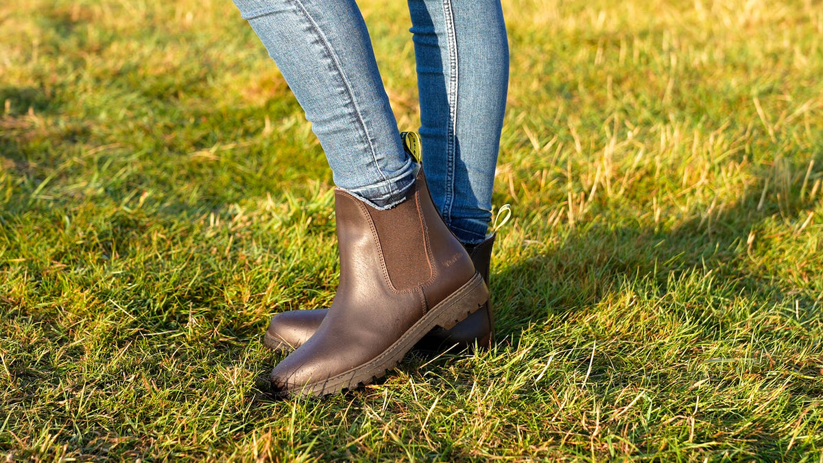 A person wearing brown leather yard boots and blue jeans standing on a grassy lawn outdoors.